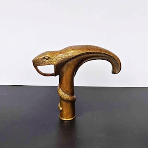 Puede incluir: Un mango de bastón de metal color bronce con forma de serpiente cobra. La cabeza de la serpiente forma la curva superior del mango, con escamas detalladas que cubren toda la superficie. Un cuerpo de serpiente más pequeño se enrosca alrededor de la base.