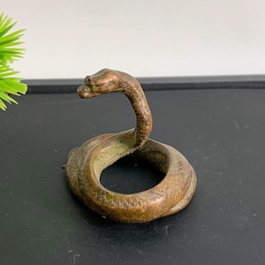 May include: A bronze snake figurine, coiled with its head raised. The sculpture has a textured surface and is set against a dark background. The snake's scales and features are detailed, showcasing a vintage aesthetic.