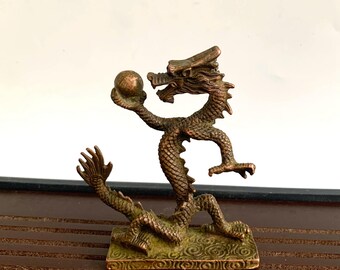 May include: A bronze dragon figurine holding a sphere. The dragon has detailed scales, a spiky mane, and outstretched claws. The base is rectangular with a swirling pattern. The figurine is on a dark brown surface.