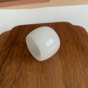 Puede incluir: Un anillo blanco y opaco con forma rectangular redondeada. El anillo está hecho de un material liso y pulido y se encuentra sobre una superficie de madera. El diseño del anillo es simple y elegante, adecuado para el uso diario.