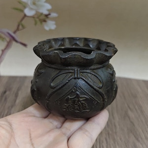 May include: A dark brown, cast metal vessel shaped like a drawstring pouch with a scalloped rim. The exterior features a molded rope design tied in a bow at the center, surrounding a square panel with embossed Chinese characters.