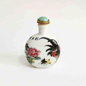 May include: A small white porcelain snuff bottle with a turquoise stone stopper. The bottle is decorated with a black and red rooster, a pink flower, and a Chinese character that reads "大吉" (pronounced "da ji"), which means "great fortune".