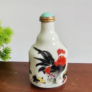 May include: A white ceramic bottle with a rooster design. The rooster has red, black, and yellow details. The bottle has a gold rim and a turquoise stopper. The bottle is likely a decorative item.