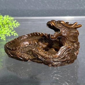 May include: A bronze-coloured dragon-shaped ashtray. The ashtray features a detailed dragon design with an open mouth and intricate scales. The dragon's body forms the ashtray's bowl, with a detailed tail and head.
