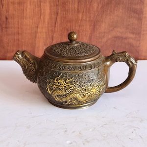 Puede incluir: Tetera de bronce con intrincados diseños de dragones y nubes. La tetera presenta una boquilla y un asa en forma de dragón, y una tapa detallada con un pequeño pomo. El diseño general es ornamentado y detallado, lo que sugiere un enfoque en la decoración de inspiración asiática.