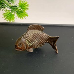 Könnte beinhalten: Eine bronzefarbene Fischfigur mit detaillierten Schuppen und Flossen. Der Fisch ist auf einer dunklen Oberfläche platziert, die sein strukturiertes Design zeigt. Die Skulptur hat eine Vintage-Ästhetik, was darauf hindeutet, dass es sich um einen Dekorationsartikel oder ein Sammlerstück handeln könnte.