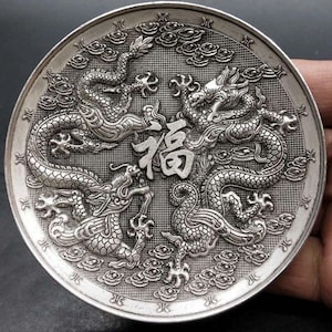 Könnte beinhalten: Eine silberne Münze mit detaillierten Reliefs von vier Drachen, die ein chinesisches Schriftzeichen umgeben. Die Münze hat einen strukturierten Hintergrund mit Wolkenmotiven und einen dekorativen Rand.