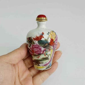 May include: A small, white ceramic bottle with a red stopper and gold trim. The bottle is decorated with a colorful floral and bird design, featuring red, pink, yellow, and green hues. The bottle is held in a hand.
