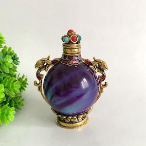 May include: A round purple and blue glass bottle with a gold-toned metal base and neck. The bottle features two gold dragon-shaped handles on the sides and a stopper adorned with small red and turquoise cabochons and intricate metal detailing.