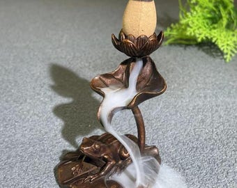 Könnte beinhalten: Bronze-Räuchergefäß mit einem Frosch auf einem Seerosenblatt und einer Lotusblume, die einen Räucherkegel hält. Der Kegel ist beige mit einer weißen Spitze, und Rauch fließt das Seerosenblatt hinunter. Es ist ein dekorativer Artikel.