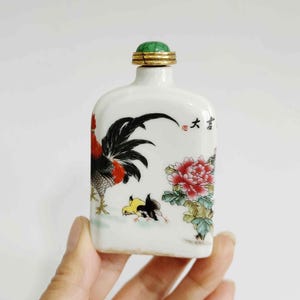 May include: A white porcelain snuff bottle with a green jade stopper. The bottle is decorated with a hand-painted design of a rooster and two chicks, along with a pink flower. The bottle has a gold rim and the Chinese character "大" is painted on the side.
