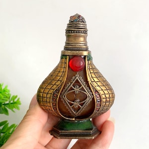 May include: A decorative metal perfume bottle featuring a textured gold-toned grid pattern, a central red cabochon stone, and intricate silver-toned filigree detailing. The bottle has a tapered neck, a small stopper, and a green-enameled base.