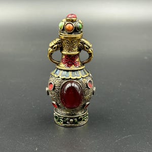 Może przedstawiać: Ozdobny, antyczny flakon perfum z bogatym wzorem. Butelka wykonana jest głównie z metalu w kolorze złota, ozdobiona czerwonymi, zielonymi i pomarańczowymi kamieniami szlachetnymi. Posiada centralny, duży, owalny czerwony kamień i ozdobną pokrywę.