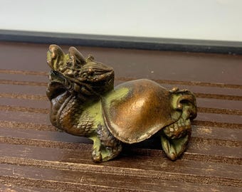 May include: A small, bronze-colored figurine of a mythical creature, possibly a dragon or a similar beast. The figurine has a detailed, textured surface with green accents, and is posed in a standing position.