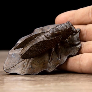 Puede incluir: Una escultura de color bronce de una cigarra posada sobre una hoja. El insecto tiene alas y segmentos corporales detallados. La hoja tiene vetas texturizadas y una forma ligeramente curvada. La escultura se sostiene en una mano sobre un fondo oscuro.