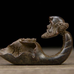 May include: A bronze-colored sculpture of a seated figure with a long beard and a lotus-shaped bowl. The figure's head is tilted, and the bowl has an inscription. The sculpture is on a wooden surface.