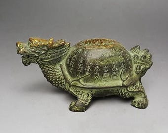 May include: A bronze-colored sculpture of a mythical creature, a hybrid of a turtle and a dragon. The sculpture has intricate details, including scales, claws, and a dragon's head with golden accents. The shell is covered in engraved text.