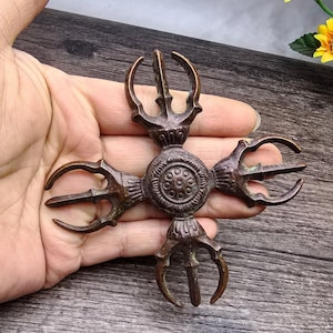 Puede incluir: Un Vajra ornamentado de color bronce, un objeto ritual con una esfera central y cuatro púas curvas que se extienden hacia afuera. El objeto tiene patrones detallados y se sostiene en una mano sobre una superficie de madera oscura.