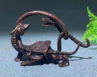 May include: A bronze-colored sculpture depicting a turtle-like creature with a snake arching over it. The turtle has detailed shell patterns and a dragon-like head. The snake's head faces the turtle. The sculpture is on a gray surface.