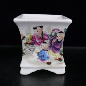 May include: A white porcelain planter with a square top and a flared body. The planter is decorated with a colorful scene of children playing, with figures in purple, pink, and blue clothing. The planter has a small, footed base.