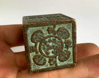 May include: A small, square, antique bronze seal with intricate carvings. The seal has a verdigris patina, indicating age. The sides feature detailed relief patterns, including circular and geometric designs. The seal is held in a hand.