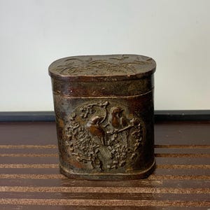 Puede incluir: Una caja rectangular marrón antigua con tapa con bisagras. La caja presenta un relieve detallado de pájaros y motivos florales. La caja está hecha de un material oscuro, posiblemente metálico, y tiene un aspecto envejecido.