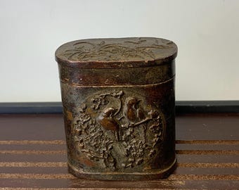 May include: A rectangular, antique brown box with a hinged lid. The box features a detailed relief of birds and floral patterns. The box is made of a dark, possibly metal material, and has an aged appearance.