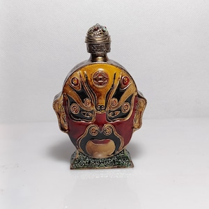 Puede incluir: Frasco de perfume antiguo y ornamentado con un diseño de rostro detallado. La botella presenta una combinación de colores rojo, amarillo y negro, con una tapa de metal decorativa. La base tiene una apariencia verde texturizada.