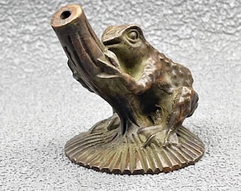 Könnte beinhalten: Bronze-Froschfigur auf einem Ast, mit einem Loch oben. Der Frosch hat eine strukturierte Haut und sitzt auf einem runden Sockel mit Blattmuster. Die Figur ist ein dekoratives Objekt, vermutlich für Haus oder Garten.