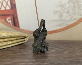May include: A bronze-colored incense holder in the shape of a frog, with its hands clasped together. The frog is positioned upside down, holding an incense stick. The background includes a wooden surface and a decorative fan.
