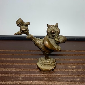 Puede incluir: Una figura de bronce que representa un oso sonriente sosteniendo una figura más pequeña. El oso está sobre una base redonda con un diseño detallado. La figura tiene un aspecto vintage y está sobre una superficie de madera oscura.