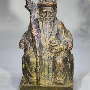 Puede incluir: Estatua de bronce de una figura anciana sentada, con barba larga y bastón. La figura lleva una túnica y una corona. La estatua está sobre una base rectangular y tiene un aspecto envejecido.