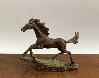 Puede incluir: Una escultura de bronce de un caballo corriendo con crin y cola ondeantes. El caballo se representa a medio paso, con las patas extendidas, sobre una base texturizada. La escultura tiene un aspecto envejecido.