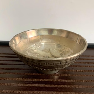 May include: A silver-colored bowl with an embossed design featuring a fish and floral elements. The bowl has a decorative border and a textured interior. The bowl is likely made of metal and is set on a dark surface.