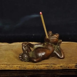 Puede incluir: Un portaincienso de rana de bronce en una pose relajada, con una varilla de incienso insertada en su cabeza. La rana descansa sobre una pila de papel envejecido, con humo que se eleva de la varilla de incienso.