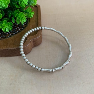 May include: A silver-toned bangle bracelet featuring a segmented design that mimics the appearance of bamboo stalks. The bracelet has a smooth, metallic finish and a circular shape, with distinct nodes spaced evenly along the entire circumference.