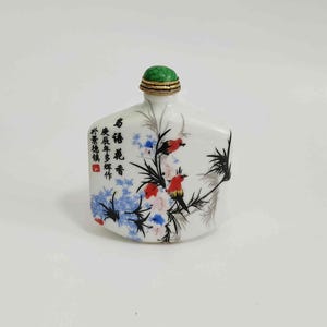 May include: A white porcelain snuff bottle with a green stopper and gold trim. The bottle is decorated with a floral and bird design in red, blue, and black, and features Chinese characters.