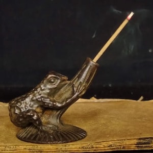 Könnte beinhalten: Ein bronzefarbener, froschförmiger Räucherstäbchenhalter. Der Frosch ist mit offenem Maul dargestellt und hält einen angezündeten Räucherstäbchen, der Rauch abgibt. Der Halter steht auf einem strukturierten Sockel, der Hintergrund ist dunkel.