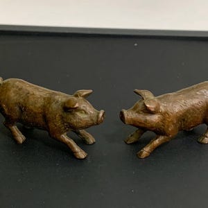 Könnte beinhalten: Zwei bronzefarbene Schweinefiguren. Die Schweine blicken sich an, ihre Schnauzen und Ohren sind sichtbar. Die Figuren stehen auf einer dunklen Oberfläche, im Hintergrund ist eine kleine grüne Pflanze.