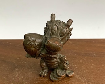 May include: A bronze-colored dragon figurine with large, expressive eyes and a whimsical expression. The dragon is carrying a sack over its shoulder and has coins at its feet. The figurine is detailed and sits on a wooden surface.