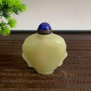 Puede incluir: Una botella de rapé de vidrio amarillo pálido con un tapón de piedra azul. La botella tiene una forma redondeada con detalles laterales sutiles. El tapón es de color azul oscuro, engastado en un metal plateado. La botella se exhibe sobre una superficie de madera oscura.