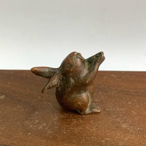 May include: A small bronze rabbit figurine with its arms raised upwards. The rabbit has long ears and a slightly weathered appearance, with hints of green patina. The sculpture is set against a neutral background.