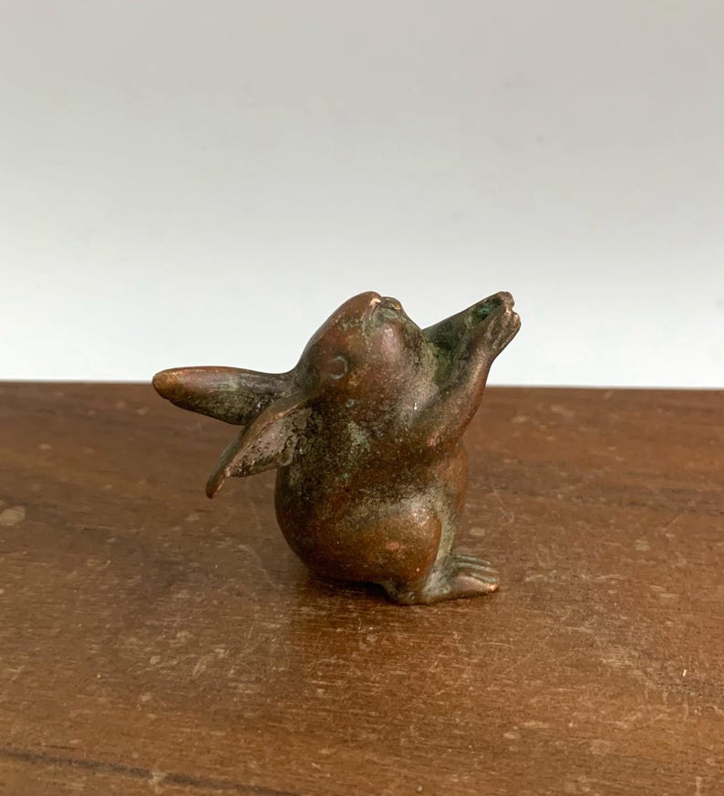May include: A small bronze rabbit figurine with its arms raised upwards. The rabbit has long ears and a slightly weathered appearance, with hints of green patina. The sculpture is set against a neutral background.