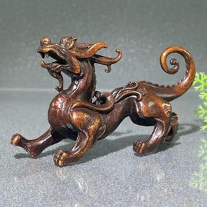 Könnte beinhalten: Bronze farbene Figur einer mythischen Kreatur mit drachenartigem Kopf, eingerolltem Schwanz und vier Beinen. Die Skulptur weist komplizierte Details auf und ist in einer dynamischen, laufenden Haltung dargestellt. Die Figur ist wahrscheinlich ein Dekorationsartikel.