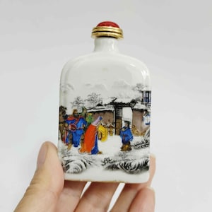 May include: A white porcelain snuff bottle with a red and gold stopper. The bottle features a painted scene of figures in colorful robes in a snowy landscape. The bottle is held in a hand.