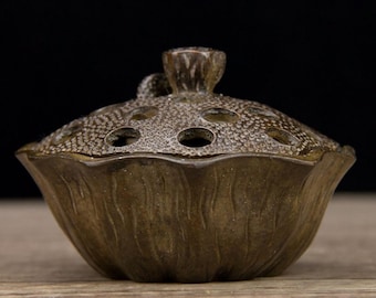May include: A bronze-colored incense burner with a lotus flower design. The lid features multiple circular openings and a textured surface. The base has a petal-like shape, and the overall design is detailed and ornate.