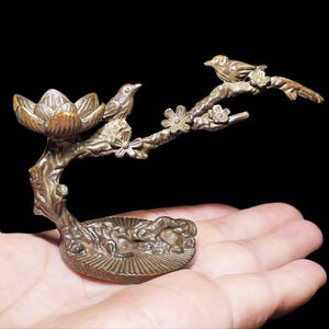 Puede incluir: Una escultura decorativa de color bronce que representa una rama con dos pájaros posados. La rama tiene flores delicadas y un elemento en forma de loto. La escultura se sostiene en una mano sobre un fondo negro.