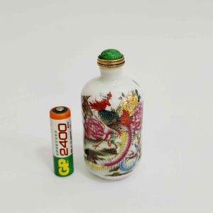 May include: A small, white ceramic bottle with a green stopper and gold trim. The bottle features a colorful painting of a phoenix and flowers. A standard AA battery is next to the bottle.