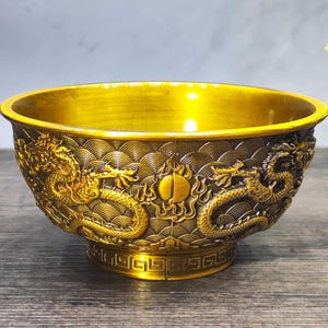 May include: A gold-colored decorative bowl with intricate dragon and wave pattern carvings. The bowl has a wide rim and a detailed base, showcasing a blend of traditional artistry and design. The bowl's surface reflects light, highlighting its metallic finish.