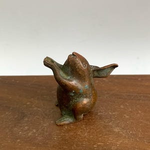 May include: A small, bronze-colored rabbit figurine with its arms raised. The rabbit has large ears and a slightly weathered appearance, with hints of green patina. The figurine is set against a wooden surface.